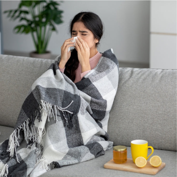 A girl sneezing on her couch and on her side is a freshly sliced lemon and a jar of mad honey